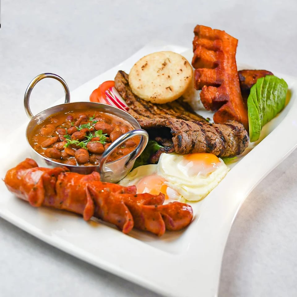 A large platter of Ecuadorian food, including grilled steak, fried eggs, beans, sausage, and avocado.