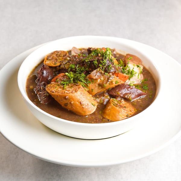 Hearty Ecuadorian stew with sausage, meat, and vegetables, garnished with fresh herbs in a white bowl.