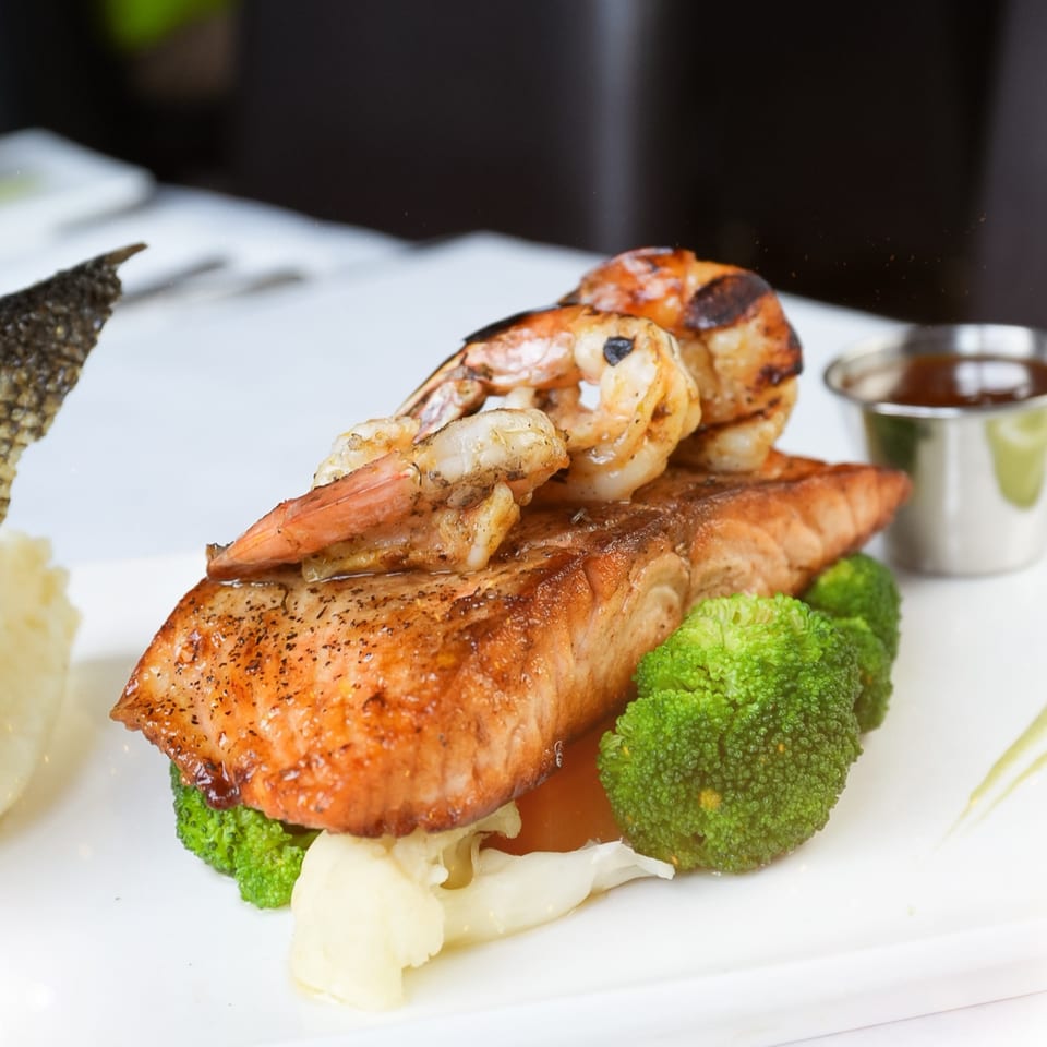 Grilled salmon and shrimp with broccoli and mashed potatoes, served with a side of sauce.